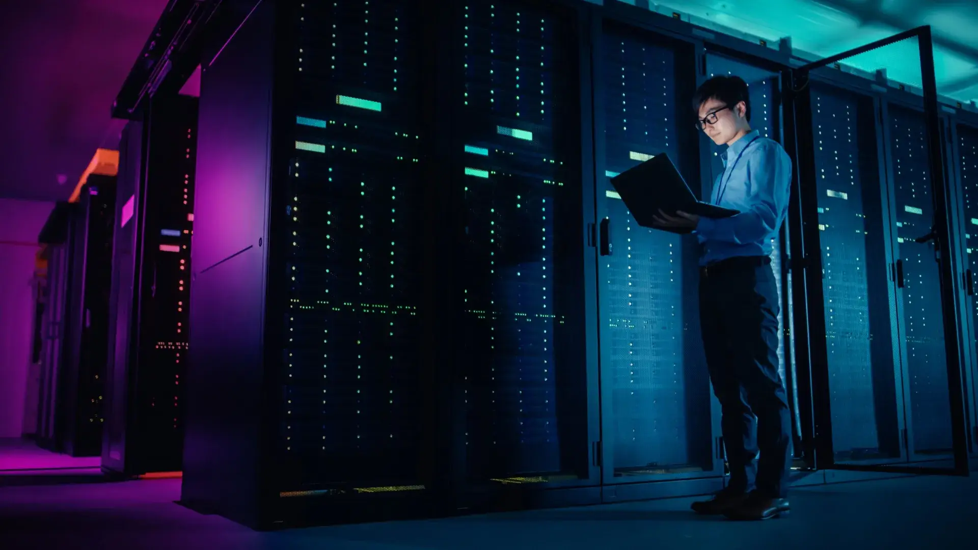 A person stands in a server room with glowing blue and purple lights, holding an open laptop, showcasing managed IT services in front of several tall racks filled with computer servers.